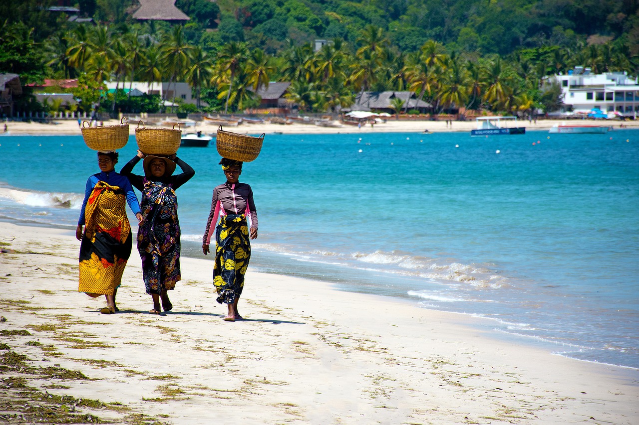 Trois femmes malgaches, vêtues de robes colorées, marchent sur une plage de sable blanc du nord de Madagascar. Chacune porte sur la tête un panier tressé