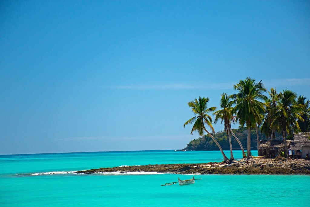 Grand ciel bleu, plage de sable blanc, mer turquoise, 5 cocotiers et des petites cabanes de plages au loin. Un petit pirogue de pécheur en premier plan. 